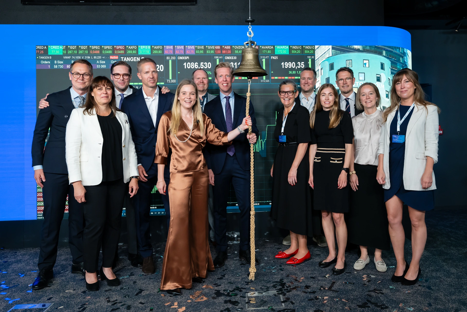 Nina Bergman och Per Andersson poserar vid börsklockan tillsammans med anställda från Tången vid noteringsceremonin för Tången B på Nasdaq. I bakgrunden syns handelsskärmar med aktiekurser.