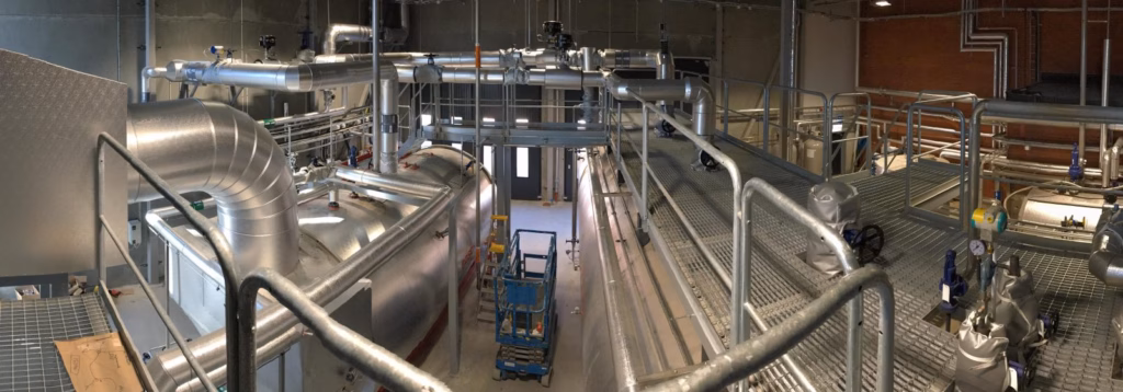 Interiör av en industriell pannanläggning med stora isolerade pannor, metallrör, ventiler och gångplattformar.